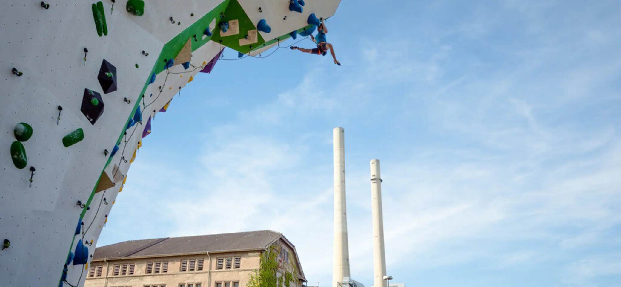 © Im Vordergrund Kletterer beim Seilklettern am Kletterturm im Außenbereich, im Hintergrund Kletterhalle