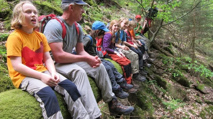 zahlreiche Kinder und ein Mann sitzen auf einem umgestürzten Baumstamm und lachen in die Kamera | © DAV Heilbronn
