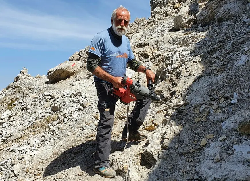 ein älterer, aber fitter und kräftiger Mann in den Bergen mit Werkzeug und Arbeitsklamotten, sowie kurzen weißen Haaren und einem Vollbart | © DAVHn