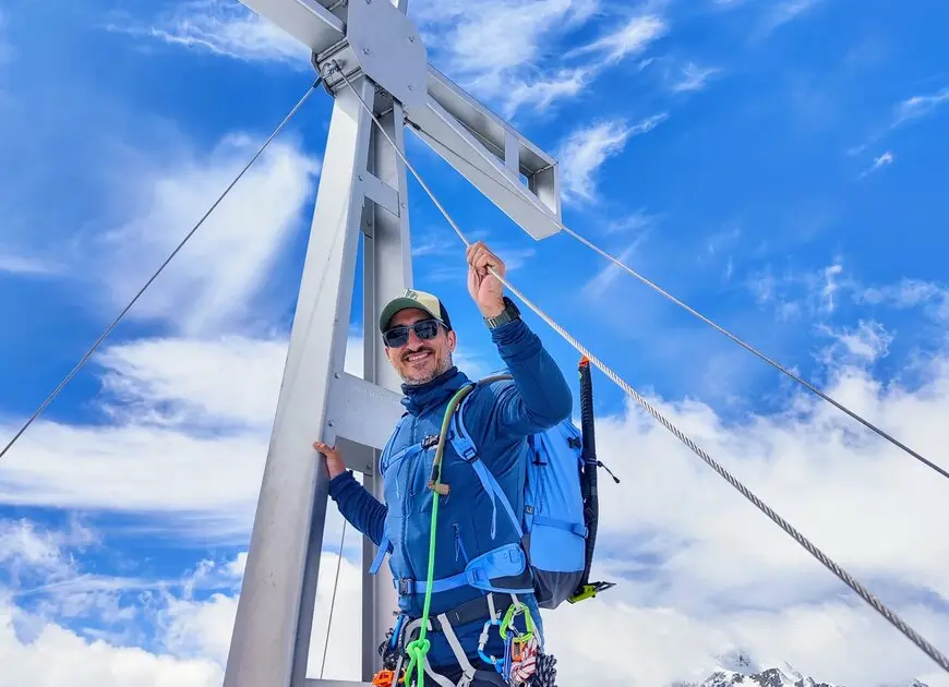 lachender Mann mit Cap und Sonnenbrille an einem Gipfelkreuz | © Özcan Keceli