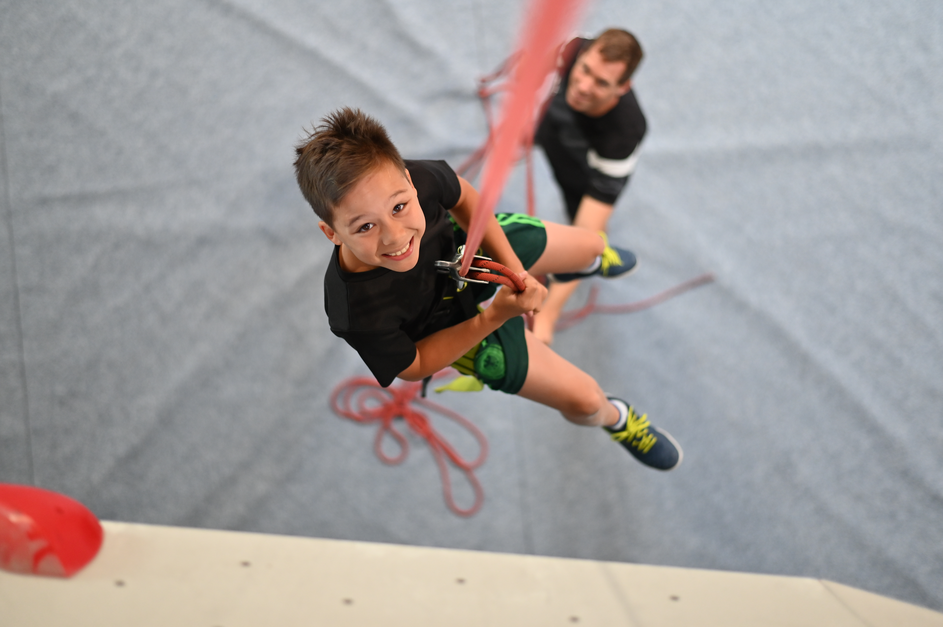DAV Heilbronn Kletterarena Indoorkurse Familienklettern | © DAV Heilbronn Bild von oben. Kinder sitzt mit Klettergurt in rotem Seil und grinst nach oben. Unten ist verschwommen ein Sicherer zu sehen. | © DAV Heilbronn